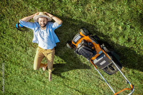 Obraz na plátně Handsome man resting on the grass near the lawn mower enjoying gardening on the