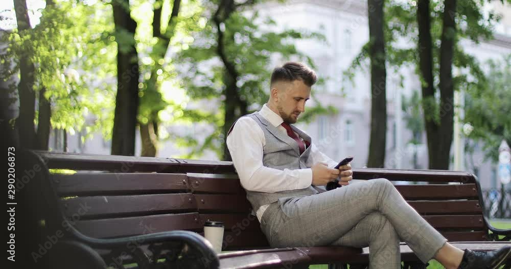 A businessman in an expensive suit sits on a park bench and reads messages on a mobile phone. A businessman in an expensive suit sits on a park bench and reads messages on a mobile phone.