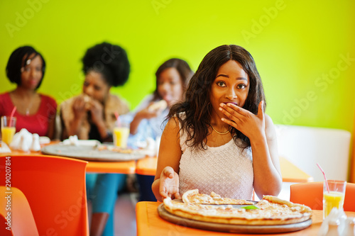 African woman with pizza sitting at restaurant against dark skinned girls. Surprised face.