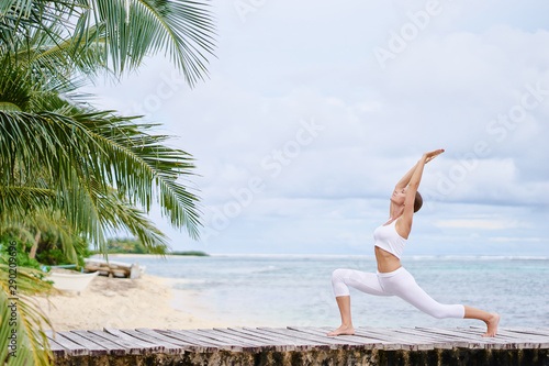 Happy young woman standing in yoga pose on wooden pier. Practicing yoga with ocean beach in the background.