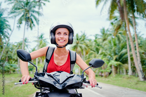 Tropical travel and transport. Happy young woman in helmet riding scooter on the road with palm trees.