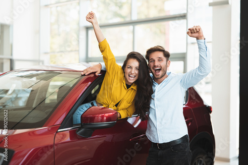 young couple looking for car in dealership center beautiful woman is sticking out the vehicle window handsome man is standing near both with hands up