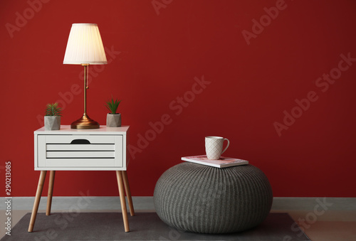 Modern table with lamp and pouf near color wall in room