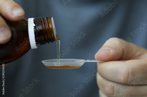 Close-up of male hands holding holding bottle of cough expectorant syrup and pouring it with measuring spoon. Ill man taking medicine. Healthcare and treatment concept. Blurred background