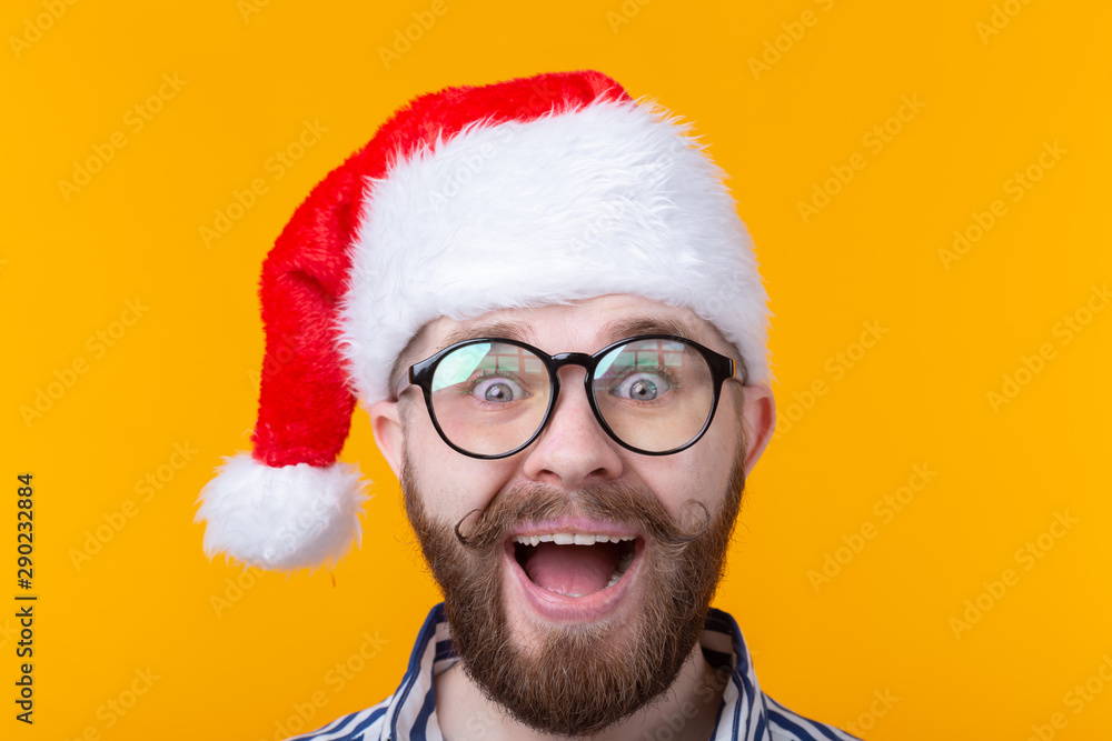 Joyful crazy cute young hipster male in red santa claus hat posing over yellow background. The concept of Christmas and the new year. Advertising space.