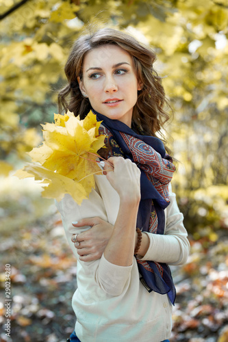 Close-up portrait of attractive brunette girl holding yellow maple leaves in her hand in park. Golden autumn foliage of trees in the background