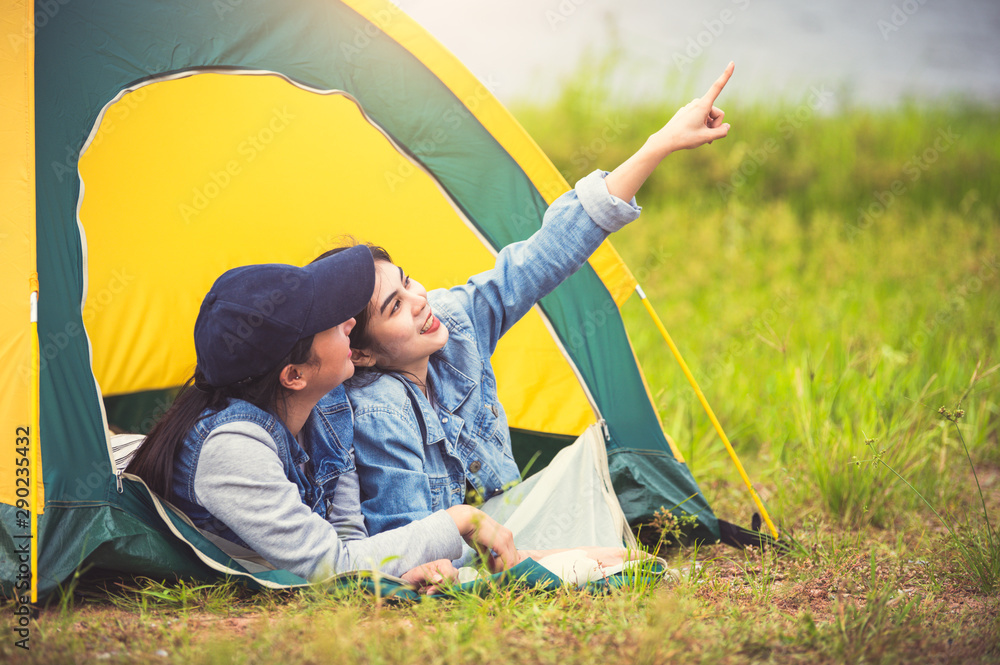 Two close friend Asian friendship relax in camping tent in green meadow on  lake side view background. Girl pointing finger to sky. People lifestyle  travel on vacation concept. Summer picnic activity Stock