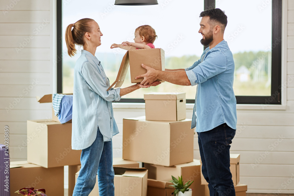 Bearded dark-haired man give daughter sit in box blonde-haired wife, they smile. Background moving box and big window Bearded dark-haired man give daughter sit in box blonde-haired wife, they smile. Background moving box and big window