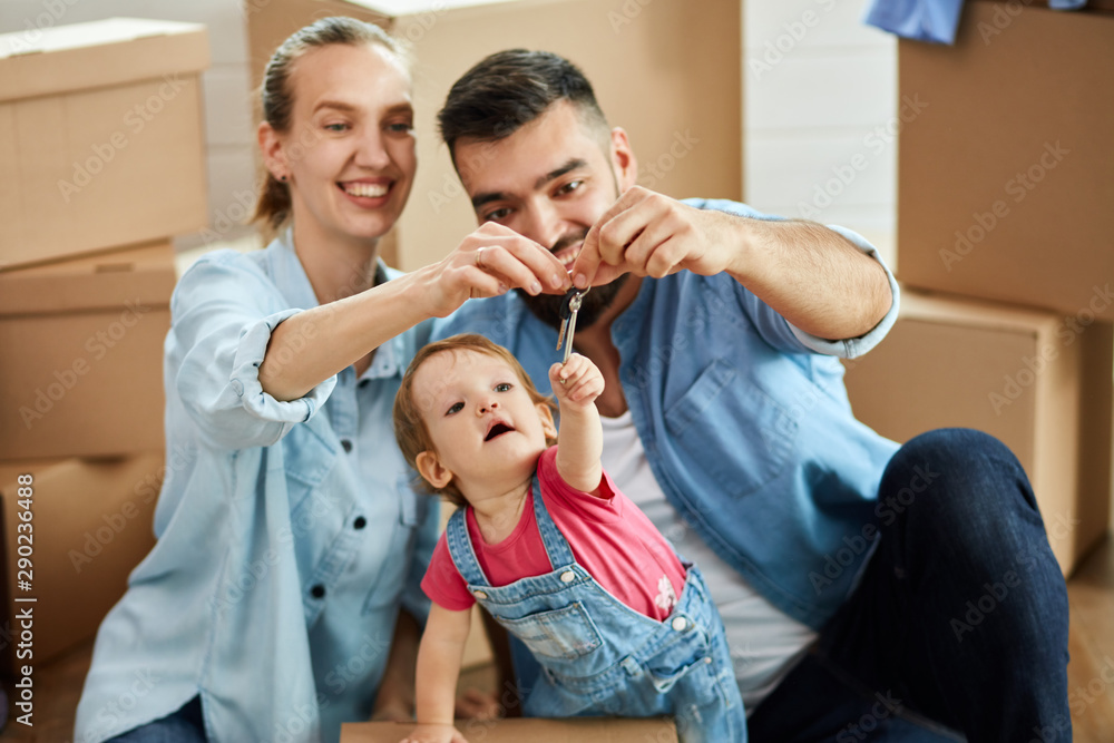 Fototapeta premium Family sit on floor near open box. Man and woman hold keys. Child want to take it. Background moving boxes