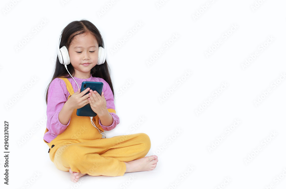 Attractive little Asian child girl in pink-yellow dungarees sitting with headphones to enjoys listening music by smartphone over white background with copy space. Attractive little Asian child girl in pink-yellow dungarees sitting with headphones to enjoys listening music by smartphone over white background with copy space.
