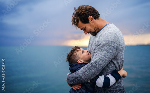 Father with small son on a walk outdoors standing on beach at dusk.