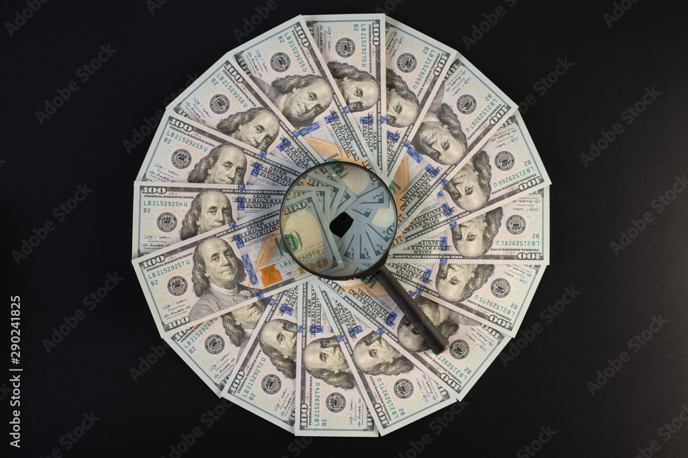 A pile of one hundred dollar bills laid out in a circle. In the middle of a magnifier, on a black background