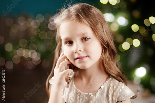 Portrait of a smiling long-haired little girl in dress on background of Christmas lights. Little girl talking on the phone. Winter holidays. Copy space