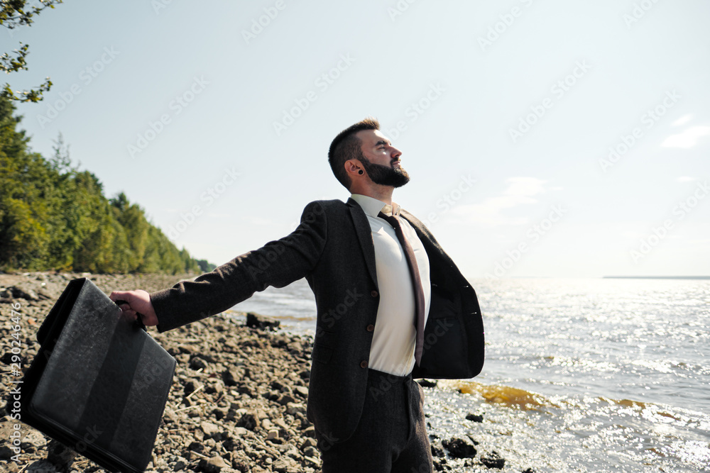 Young attractive male businessman in a brown suit with a briefcase stands on the seashore. Unfastens shirt, loosens tie and raises hands to sea.