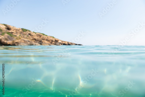 Turquoise blue sea above water surface and underwater. Sun glare at the bottom of ocean. Waves underwater and rays of sunlight shining through. Transparent water and light at sand.