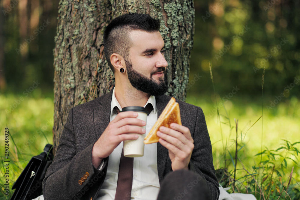 Young attractive business man with beard sitting on green grass under tree and resting in park. Drinks coffee and eats a sandwich. Relaxation, tired of work, lunch break outdoor. Young attractive business man with beard sitting on green grass under tree and resting in park. Drinks coffee and eats a sandwich. Relaxation, tired of work, lunch break outdoor.