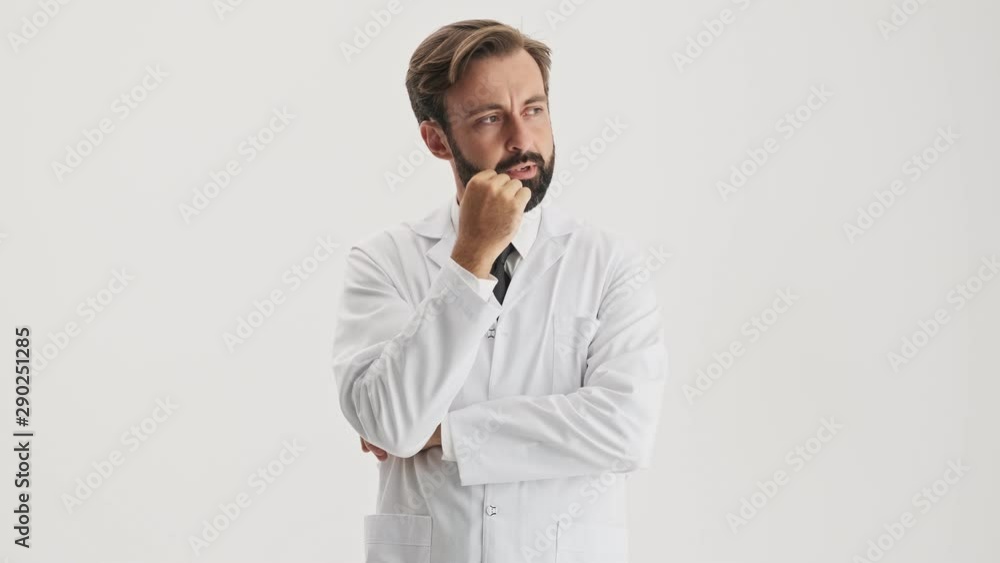 Thoughtful young bearded man doctor in white professional coat touching his nose bridge with hand and looking to the side over gray background isolated Thoughtful young bearded man doctor in white professional coat touching his nose bridge with hand and looking to the side over gray background isolated