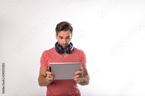 Young beautiful hipster guy with headphones on neck using digital gadget while standing isolated in studio. Man watching online video on touch pad