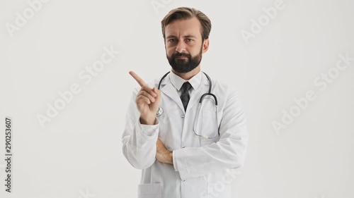 Angry young bearded man doctor in white professional coat with stethoscope saying no with waving finger and shaking his head negatively while looking at the camera over gray background isolated