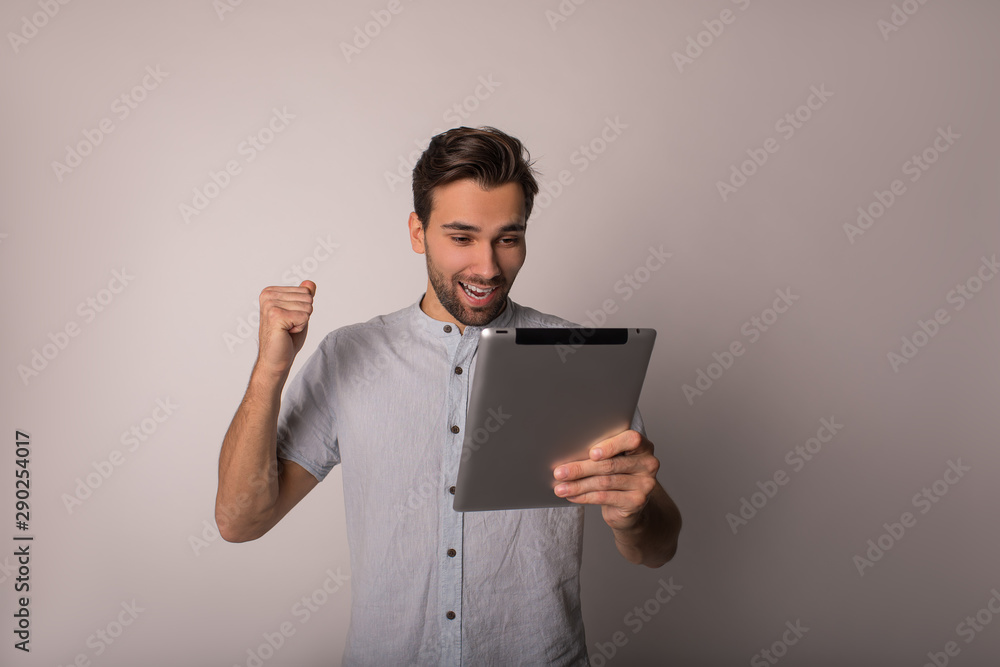 Happy smiling man university student reading good news on website via digital gadget while standing isolated in studio. Celebrating victory Happy smiling man university student reading good news on website via digital gadget while standing isolated in studio. Celebrating victory