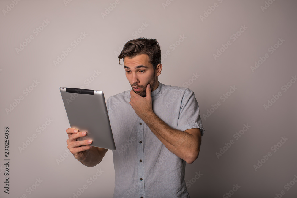 Young astonished man in office shirt received shocked news in e-mail on portable touch pad while standing isolated in studio against wall. Young astonished man in office shirt received shocked news in e-mail on portable touch pad while standing isolated in studio against wall.