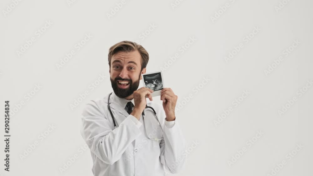 Excited young bearded man doctor in white professional coat with stethoscope showing ultrasound scan of his baby over gray background isolated Excited young bearded man doctor in white professional coat with stethoscope showing ultrasound scan of his baby over gray background isolated