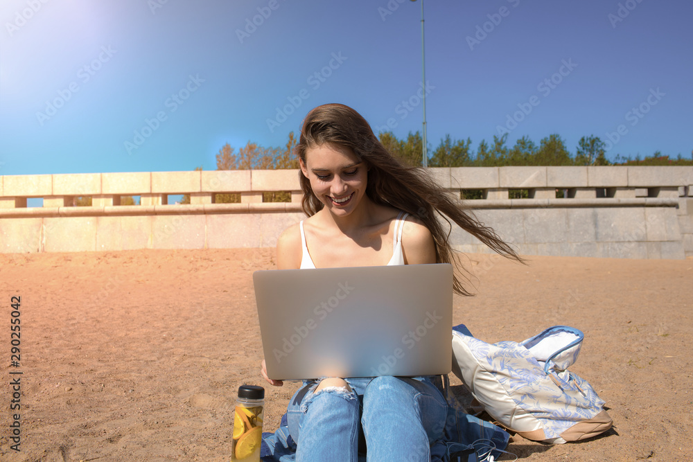 Happy smiling woman freelance copywriter typing text on laptop computer while relaxing outdoors on the beach in sunny day during summer vacation. Cheerful hipster girl keyboarding on netbook