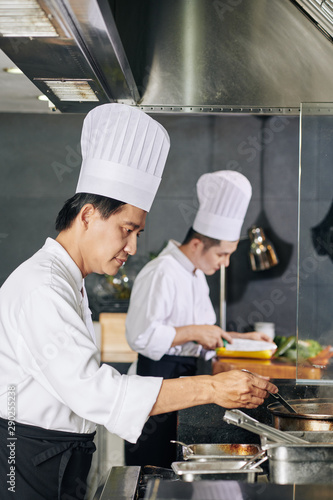 Asian mature chef in white hat standing near the stove and preparing food together with his assistant in the background in the kitchen