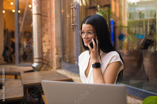 Fashionable dark hair caucasian female in glasses skilled freelance worker talking via mobile phone while sitting outside coffee shop. Woman owner having cellphone conversation after webinar