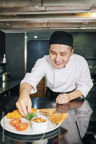 Asian young chef decorating his prepared dish with greens and smiling in the kitchen of the restaurant