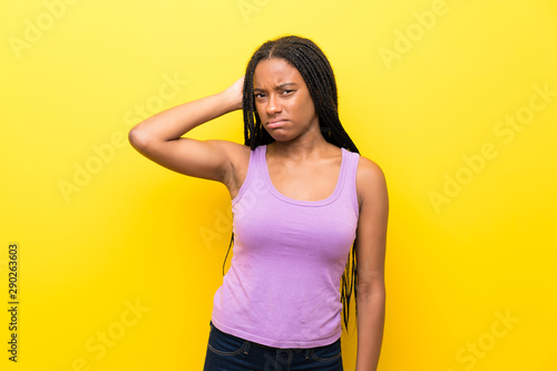 African American teenager girl with long braided hair over isolated yellow wall having doubts