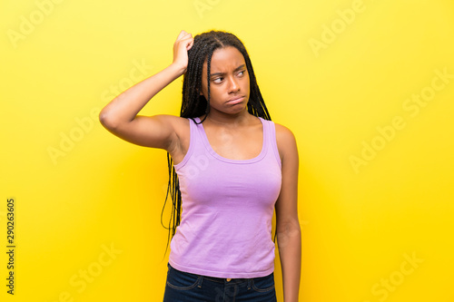 African American teenager girl with long braided hair over isolated yellow wall having doubts while scratching head