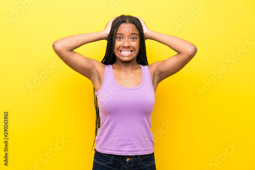 African American teenager girl with long braided hair over isolated yellow wall frustrated and takes hands on head