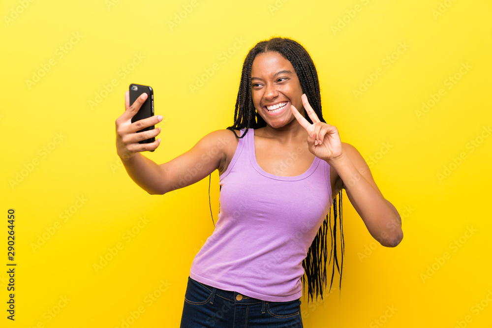 African American teenager girl with long braided hair over isolated yellow wall making a selfie African American teenager girl with long braided hair over isolated yellow wall making a selfie