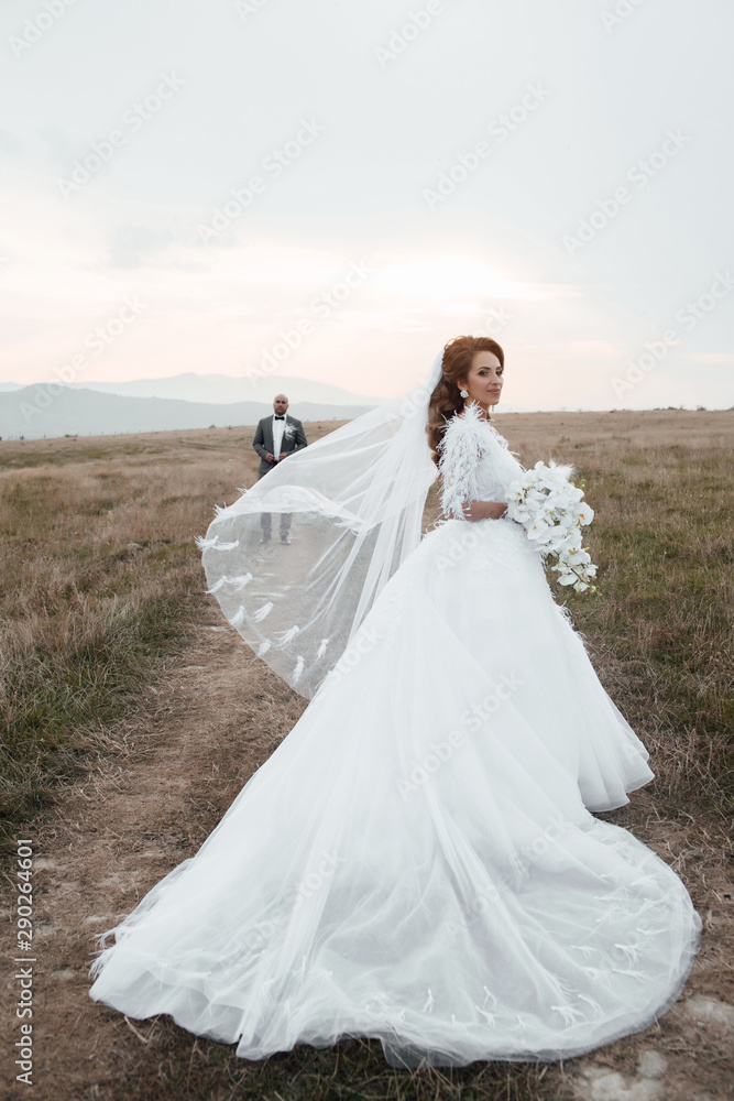Woman in fashionable dress waving veil wind. Stylish and beautiful. Bridal bouquet of orchids.  Loving wedding couple outdoor. Beautiful woman with professional make up and hairstyle.