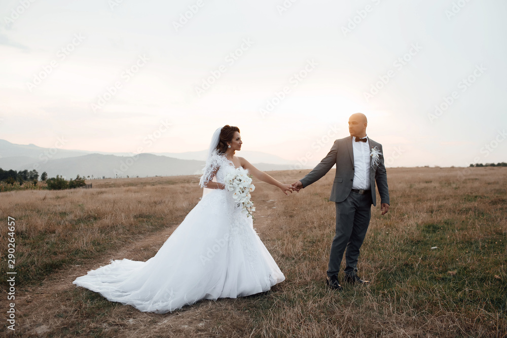 Romantic wedding couple having fun together outdoor at field. Groom and bride in a wedding dress going through the field on a background of blue sky at sunset. Wedding. Loving wedding couple