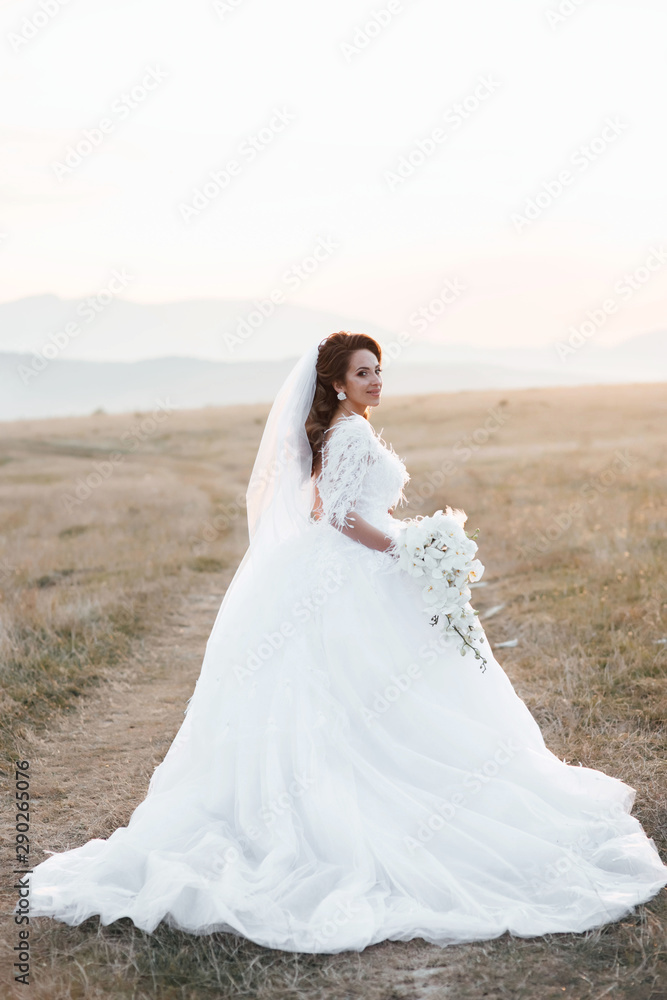 Happy bride in wedding dress posing on the road in a field at sunset.   Beautiful young bride is holding a wonderful bouquet of orchid.  Beautiful woman with professional make up and hairstyle.