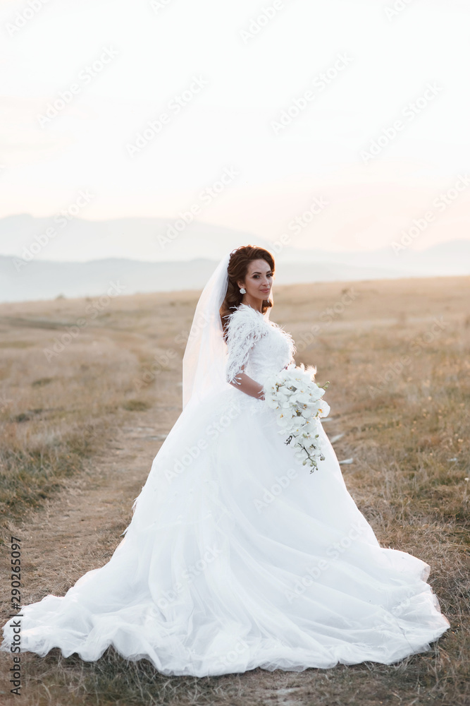Happy bride in wedding dress posing on the road in a field at sunset.   Beautiful young bride is holding a wonderful bouquet of orchid.  Beautiful woman with professional make up and hairstyle.