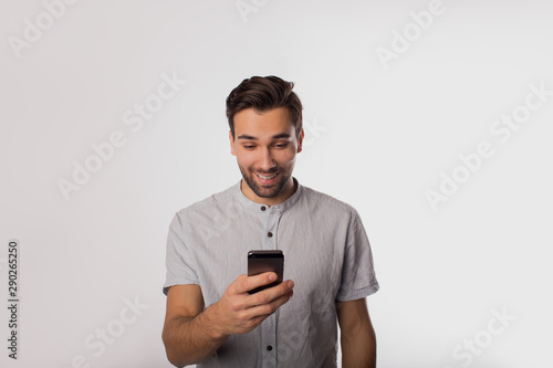 Happy smiling man in casual wear watching video on mobile phone while standing isolated in studio against wall with copy space for promotional content. Joyful male chatting on cellphone © ZoFot
