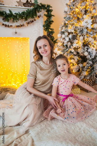 Merry Christmas and Happy Holidays. Cheerful mom and her cute daughter sitting on a plaid near a fireplace decorated with a golden garland and a beautiful Christmas tree