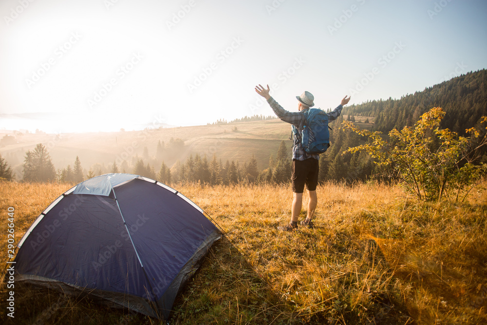 Happy hiker man meet sunset on mountain top. Tourist admire inspirational landscape with hands up. Climber reached mountain peak