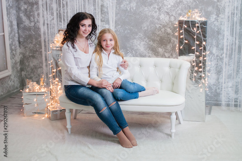 Two very pretty girls, sisters celebrates Christmas, new year. Girls in the New Year's interior of the room are sitting on a white sofa against the background of garlands