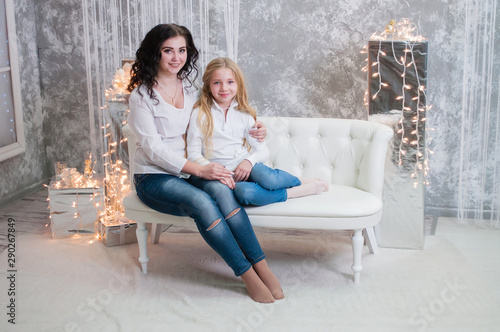 Two very pretty girls, sisters celebrates Christmas, new year. Girls in the New Year's interior of the room are sitting on a white sofa against the background of garlands