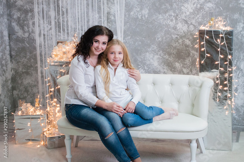 Two very pretty girls, sisters celebrates Christmas, new year. Girls in the New Year's interior of the room are sitting on a white sofa against the background of garlands