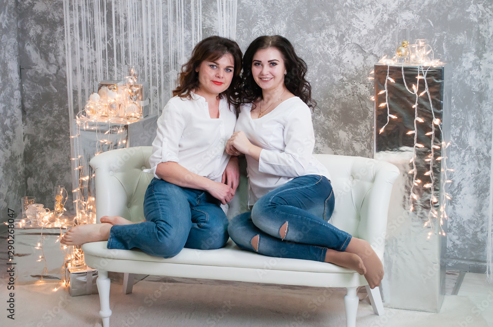 Two very pretty young women celebrate Christmas, the new year. Girls in the New Year's interior of the room are sitting on a white sofa against the background of garlands Two very pretty young women celebrate Christmas, the new year. Girls in the New Year's interior of the room are sitting on a white sofa against the background of garlands