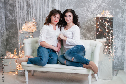 Two very pretty young women celebrate Christmas, the new year. Girls in the New Year's interior of the room are sitting on a white sofa against the background of garlands