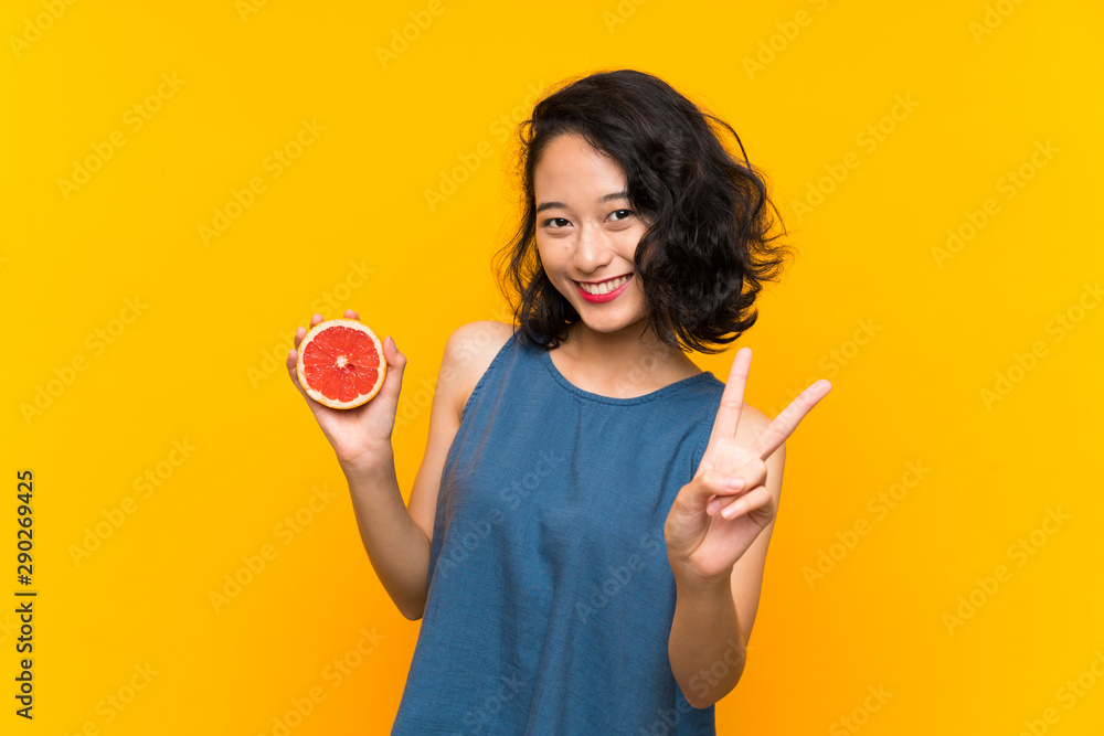 Young asian girl holding a grapefruit over isolated orange background smiling and showing victory sign Young asian girl holding a grapefruit over isolated orange background smiling and showing victory sign