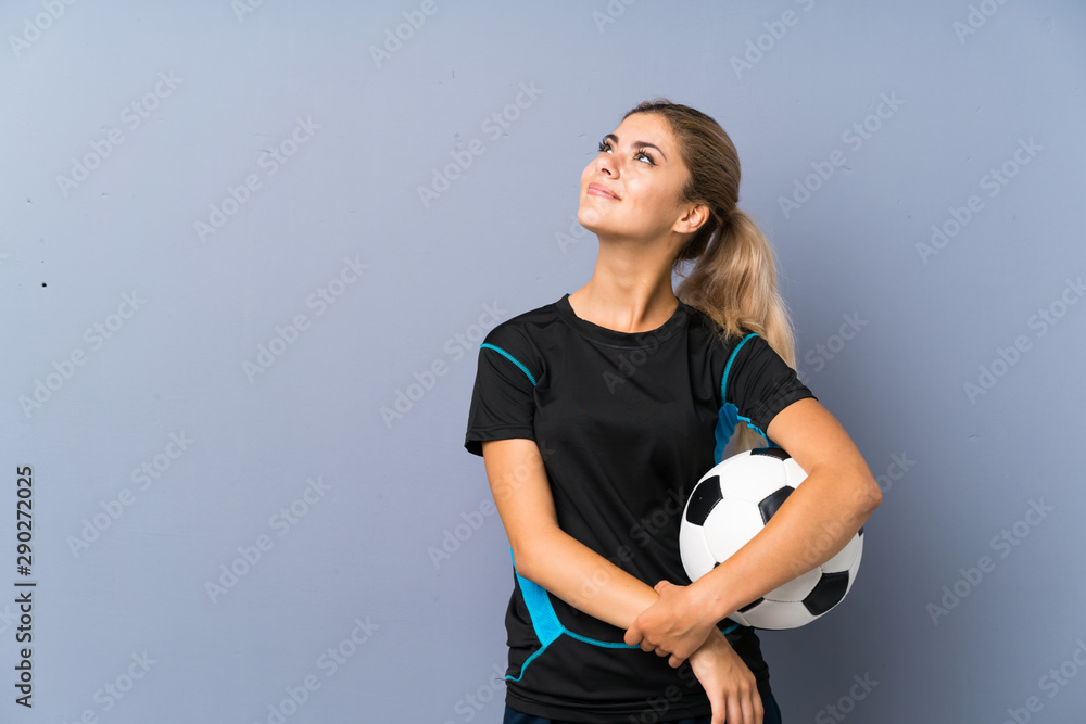 Blonde football player teenager girl over grey wall looking up while smiling Blonde football player teenager girl over grey wall looking up while smiling