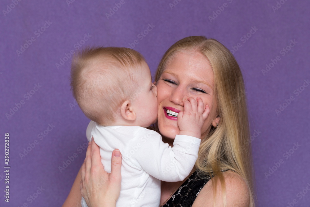 Medium horizontal portrait of adorable happy baby boy kissing his pretty smiling young blond mother holding him against mauve background Medium horizontal portrait of adorable happy baby boy kissing his pretty smiling young blond mother holding him against mauve background