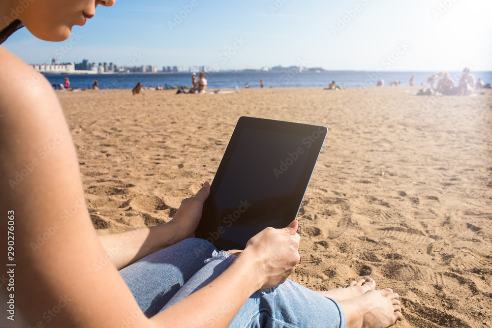 Female traveler online booking purchase via touch pad while relaxing on the beach during summer travel abroad. Hipster girl reading article via digital gadget while resting outdoors